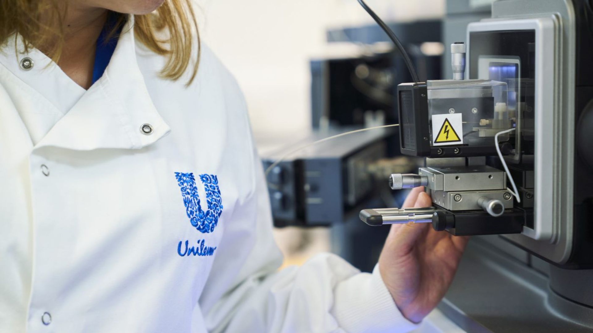 Nahaufnahme eines Wissenschaftlers in einem weißen Laborkittel mit dem Logo von Unilever, der durch ein großes digitales Mikroskop schaut.